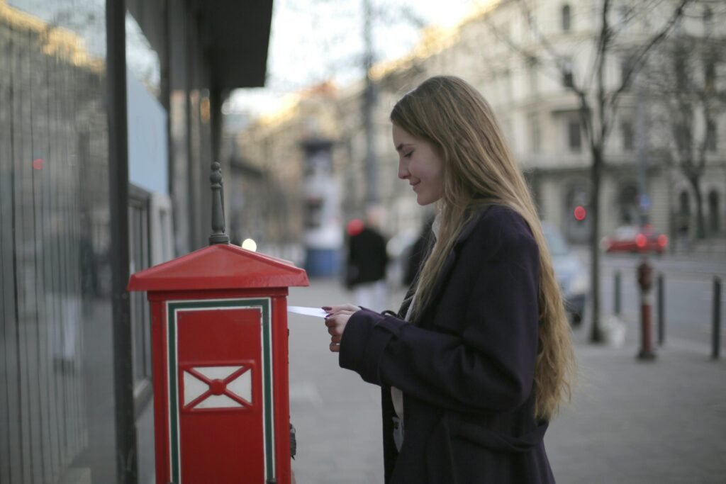 A woman wearing a coat posts a letter in a red postbox on a city street.